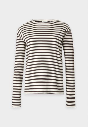 Femme debout portant une chemise rayée noire et blanche à manches longues, un jean bleu foncé à jambes larges et des chaussures marron, une main dans la poche.