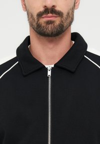 Άνδρας με σκούρο μούσι που φορεί μαύρο μπουφάν με φερμουάρ και λευκές ρίγες στους ώμους, μπροστά σε ανοιχτό ανοιχτόχρωμο φόντο.