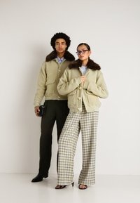 Két modell bézs színű bunda dzsekiket visel, sötét szőrme gallérral; az egyik kockás széles nadrágban és szandálban, a másik sötét csíkos nadrágban és öltönycipőben.