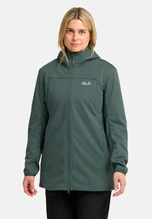 Donna che indossa una giacca verde con cappuccio Jack Wolfskin con cerniera completa frontale e tasche, in piedi davanti a uno sfondo semplice.