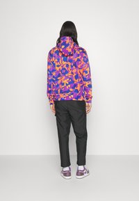 Barevná mikina s abstraktním vzorem v odstínech fialové a oranžové, spárovaná s černými kalhotami a teniskami s fialovými akcenty. Měkká tkanina, volný střih.