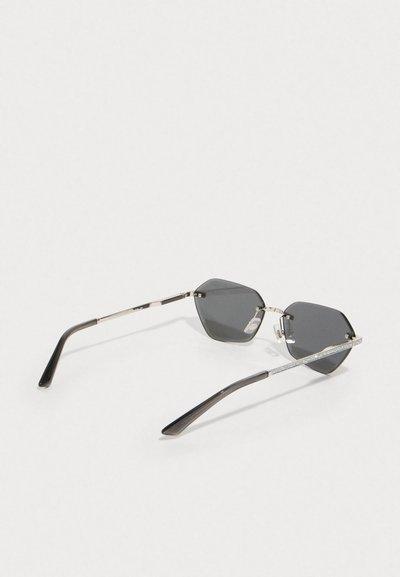 Lunettes de soleil hexagonales avec verres noirs, monture en métal argenté, accents texturés en argent sur les branches, et bras en plastique noir brillant.