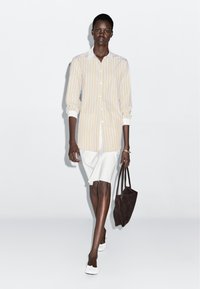 Striped long-sleeve shirt in yellow and white, paired with a white skirt. Model carries a dark brown suede bag, wearing white slip-on shoes.