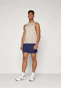 Canotta in tessuto beige chiaro con scollatura rotonda, abbinata a pantaloni shorts blu navy. Sneakers bianche con dettagli neri. Sfondo semplice.