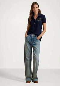 Ung kvinna som bär en marinblå pikétröja med broderad design, vida blekta jeans och bruna skor, stående med händerna i fickorna.