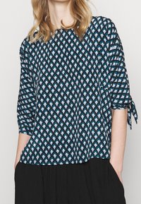 Blouse bleue à motifs avec un design de vagues dans des tons de sarcelle et de blanc, avec des manches trois-quarts et des liens aux poignets.