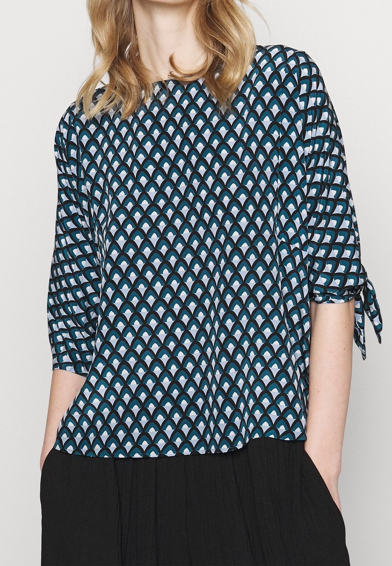 Blouse bleue à motifs avec un design de vagues dans des tons de sarcelle et de blanc, avec des manches trois-quarts et des liens aux poignets.