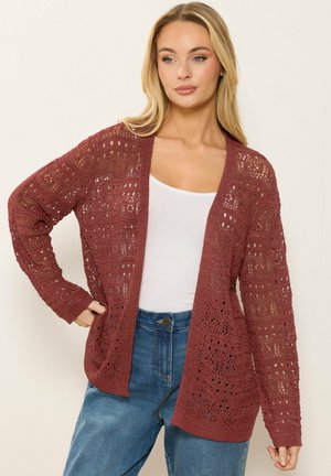 Donna che indossa un cardigan all'uncinetto color ruggine aperto sul davanti, canotta bianca e jeans blu, posando con una mano sul fianco contro uno sfondo semplice.