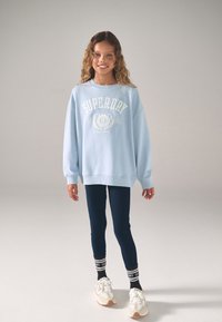 Šviesiai mėlynas itin laisvo stiliaus džemperis su baltu „SUPERDRY“ logotipu, derinamas su tamsiai mėlynomis tamprėmis ir baltais sportbačiais su tekstūruotomis padais bei juodais akcentais.