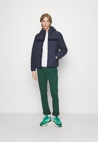 Giacca imbottita blu navy con cappuccio, caratterizzata da cuciture orizzontali e una striscia nera. Abbinata a pantaloni verdi e sneaker verdi. T-shirt bianca sotto.