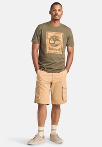T-shirt verde oliva con grafica di un albero e testo "Timberland", abbinata a pantaloni cargo beige con tasche laterali e sneakers beige.