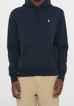 Mand iført en marineblå hættetrøje med en hvid polo-spillerlogo og beige bukser mod en ensfarvet baggrund.