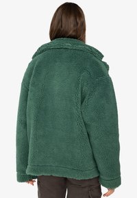 Grön fleecejacka med en strukturert yta, bred krage och avslappnad passform. Har neddragen axlar och tjocka manschetter.