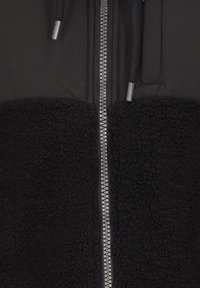 Capa exterior negra con textura de forro polar en la parte inferior, cremallera plateada en el centro y cordones en el cuello. Diseño minimalista.