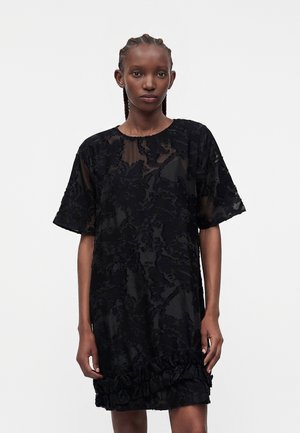 Νέα γυναίκα με κοτσιδάκια που φοράει μαύρο κοντομάνικο φόρεμα με υφασματικό λουλουδάτο σχέδιο και βολάν στο τελείωμα, στέκεται μπροστά σε λευκό φόντο.