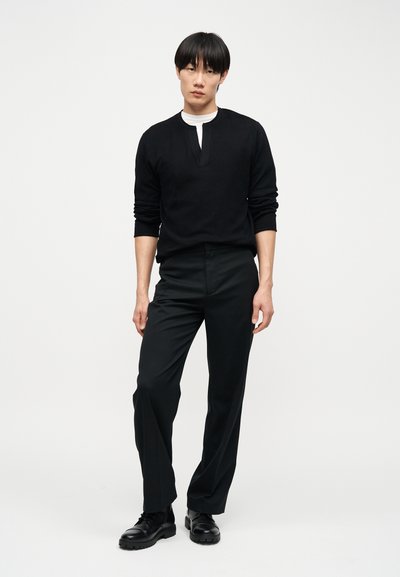 Νέος άνδρας όρθιος, φορά μαύρο μακρυμάνικο πουκάμισο με λευκό εσώρουχο, μαύρο παντελόνι και μαύρα παπούτσια σε απλό λευκό φόντο.