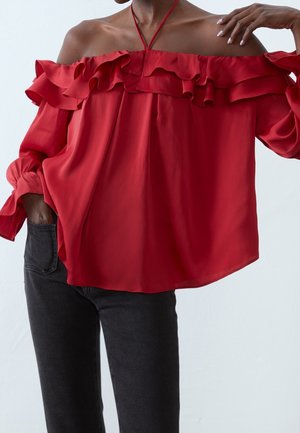 Blouse rouge à épaules dénudées avec détail à volants, associée à un jean noir, main posée sur le cou, posture détendue sur un fond uni.