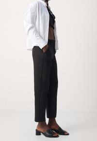 Weiße Bluse über einem schwarzen Crop-Top, kombiniert mit hoch taillierten schwarzen Hosen und schwarzen offenen Absatzschuhen. Minimalistisches Design.