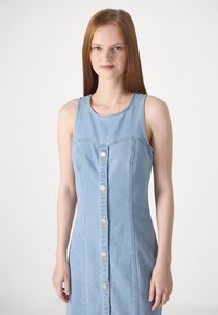 Lys blå denimkjole med en tettsittende silhuett, uten ermer og med knapplukking foran. Har subtile sømmer og rund halslinning.