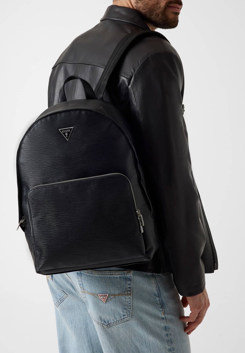 Homme portant une veste en cuir noire et un jean bleu, portant un sac à dos Guess noir texturé avec une poche avant zippée.