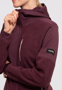 Veste en polaire bordeaux avec capuche ajustée, poche poitrine zippée et un patch logo sur la manche. Texture lisse, design structuré.