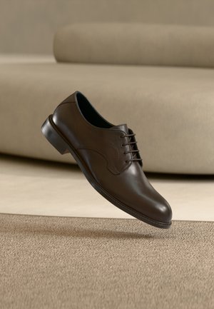 Brauner Lederschnürschuh für feierliche Anlässe schwebt über beigem Teppich, im Hintergrund ein hellbeiges Sofa.