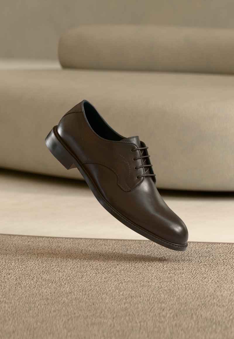 Καφέ δερμάτινο επίσημο παπούτσι με κορδόνια που αιωρείται πάνω από μπεζ χαλί με ανοιχτό καφέ καναπέ στο παρασκήνιο.