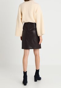 Cremfarbener gerippter Pullover mit weiten Ärmeln, kombiniert mit einem dunkelbraunen Leder-Minirock und schwarzen Ankle-Boots mit Blockabsatz.