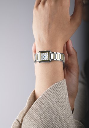 Montre-bracelet argentée et dorée avec un cadran rectangulaire, des chiffres romains et un bracelet en métal, portée sur un poignet clair.