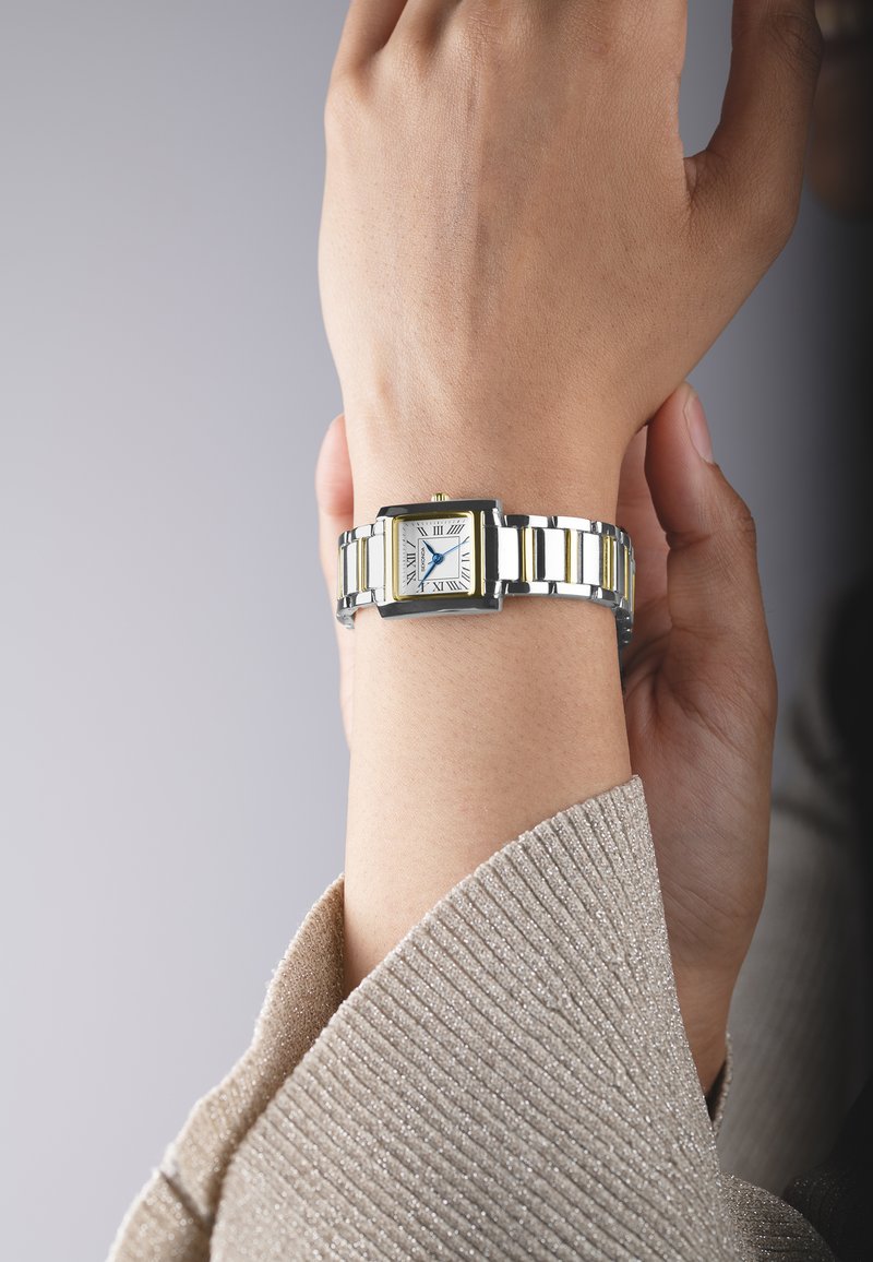 Montre-bracelet argentée et dorée avec un cadran rectangulaire, des chiffres romains et un bracelet en métal, portée sur un poignet clair.