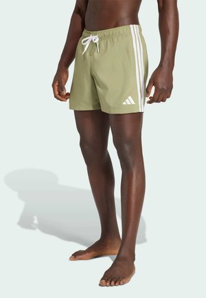 Uomo con pantaloncini da bagno Adidas verde chiaro con strisce bianche e logo, in piedi a piedi nudi su sfondo chiaro.
