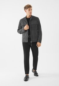 Giacca in suede grigia con chiusura a zip, due tasche sul petto e colletto alla coreana, abbinata a una camicia nera e pantaloni neri slim. Mocassini neri.
