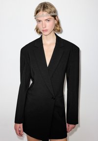 Schwarzes tailliertes Blazerkleid mit markanten Schultern, strukturierter Silhouette, tiefem V-Ausschnitt, einem Knopf und glatter Textur.