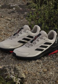 Jasnoszare buty trekkingowe z czarnymi akcentami, teksturowaną siateczką u góry i czerwoną gumową podeszwą. Posiadają logo na języku i sznurowadłach.