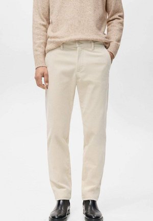 Beige Cordhosen getragen mit einem hellbraunen Pullover und schwarzen Leder-Stiefeletten an einer Person, die vor einem weißen Hintergrund steht.