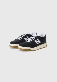 Sorte semskede sneakers med hvite lisser, med et fremtredende hvitt "N"-logo og en beige gummisåle, designet med lav profil.