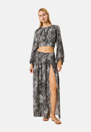 Donna con un crop top a maniche lunghe grigio e nero con stampa animalier e gonna maxi abbinata con spacco alto, con sandali, in piedi su sfondo bianco.