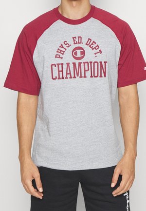 Muž nosící šedé a bordó raglánové tričko s nápisem "PHYS. ED. DEPT. CHAMPION" a logem na hrudi, v kombinaci s černými kalhotami.