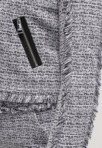 Svart och vit tweedtyg med en strukturerad mönstring; har en svart dragkedjeficka och fransdetaljer längs kanterna.