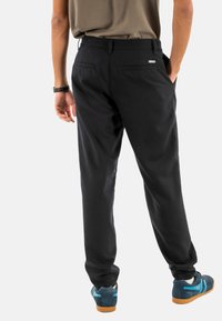 Uomo con pantaloni neri, scarpe da ginnastica blu e una camicia marrone, con una mano in tasca, visto di spalle su uno sfondo bianco.