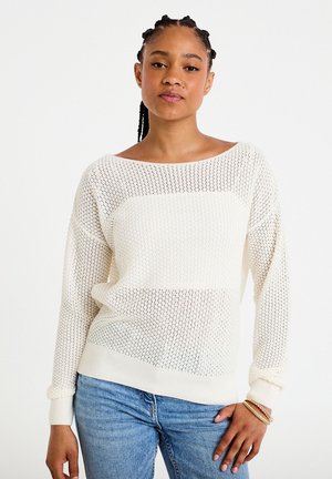 Jonge vrouw met gevlochten haar draagt een witte gebreide trui en blauwe spijkerbroek, staand met één hand op haar heup tegen een witte achtergrond.