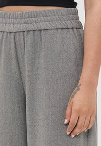 Šedé joggers s texturou a elastickým pasem. Detailní záběr ukazuje ruku s tetováním položenou na tkanině.