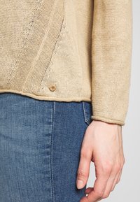 Beige gebreide trui met een gestructureerde afwerking en een decoratieve houten knoop aan de zoom, gecombineerd met blauwe denim jeans.