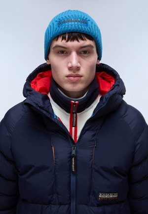 Giacca puffer blu navy con zip sul davanti, dettagli arancioni e logo ricamato. Sovrapposta a un scaldacollo in pile rosso e bianco e a un cappellino di maglia blu.