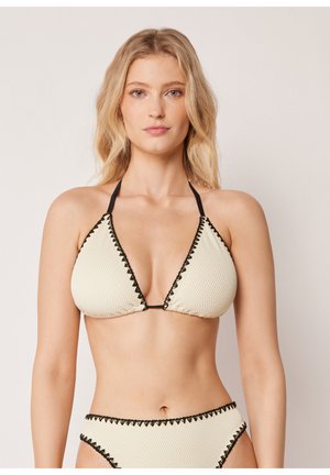 Blondynka w kremowym teksturowanym bikini z czarnym obszyciem, pozująca na jednolitym neutralnym tle.