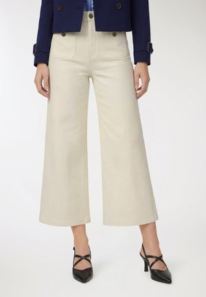 Donna con pantaloni beige a gamba larga e corti, giacca blu navy con dettagli a fibbia e décolleté nere con cinturini e punta appuntita.