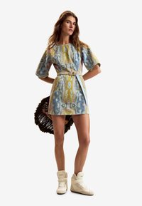 Haljina od trapera s kratkim rukavima s apstraktnim pastelnim uzorkom, strukirana remenom i ušivenim džepovima. Uparena s bež visokim tenisicama.