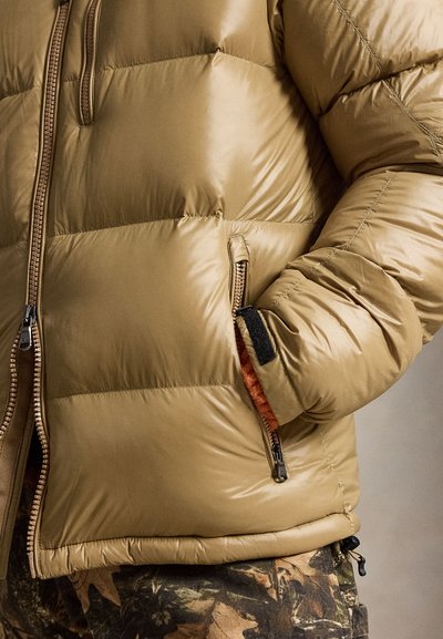 Doudoune beige avec poches zippées à l’avant et sur les côtés, une main dans la poche, associée à un pantalon camouflage.