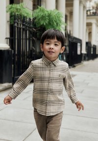 Bambino con camicia a quadri chiara con tonalità marroni, abbinata a pantaloni di velluto a coste color marrone chiaro, che cammina su un sentiero di pietra.