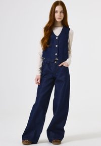 Marineblauwe dennenvest met vijf knopen over een lichtbeige lange mouw top, gecombineerd met losse marineblauwe denim brede pijpenbroek.